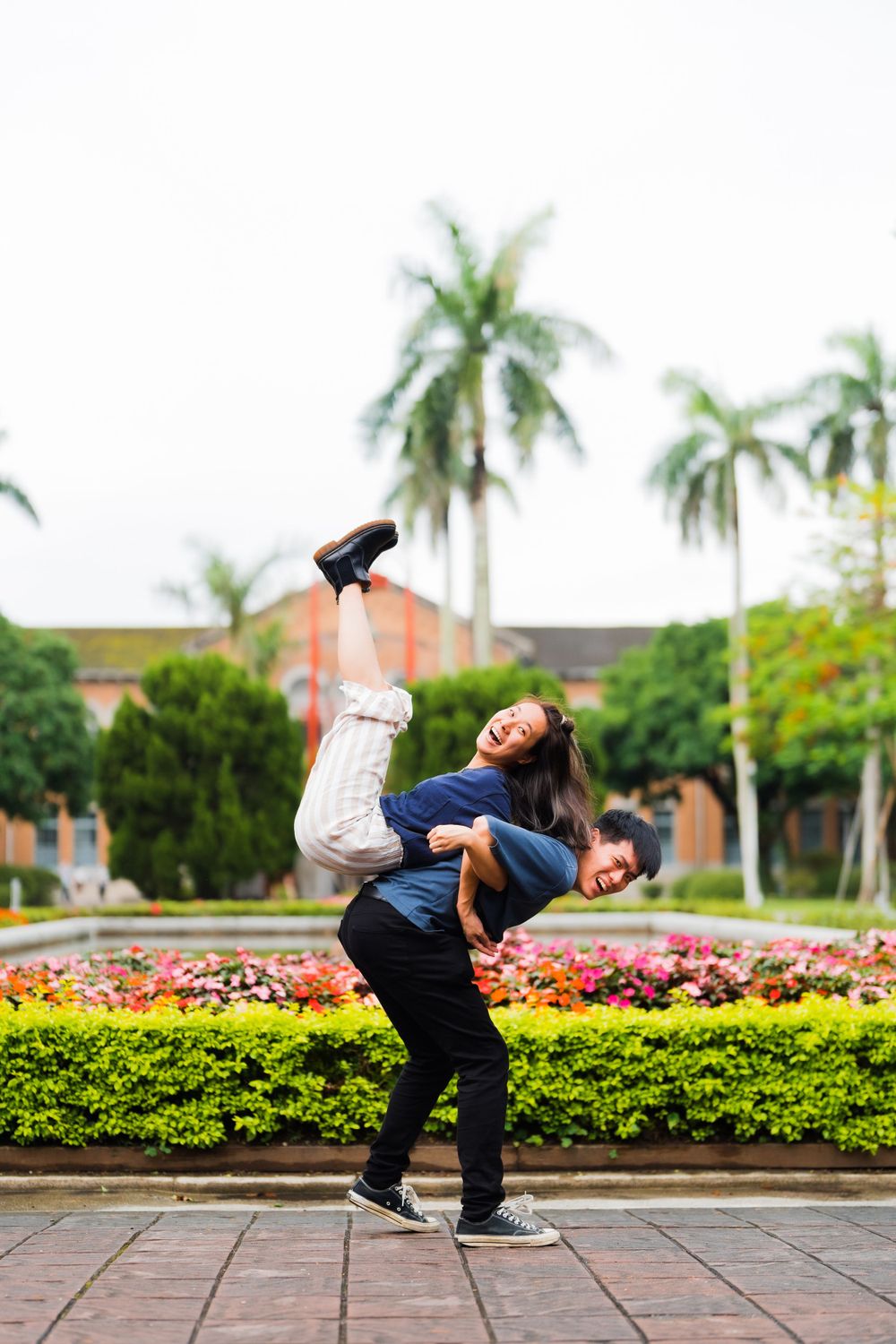 Two friends perform a playful acrobatic pose in front of a garden with palm trees.