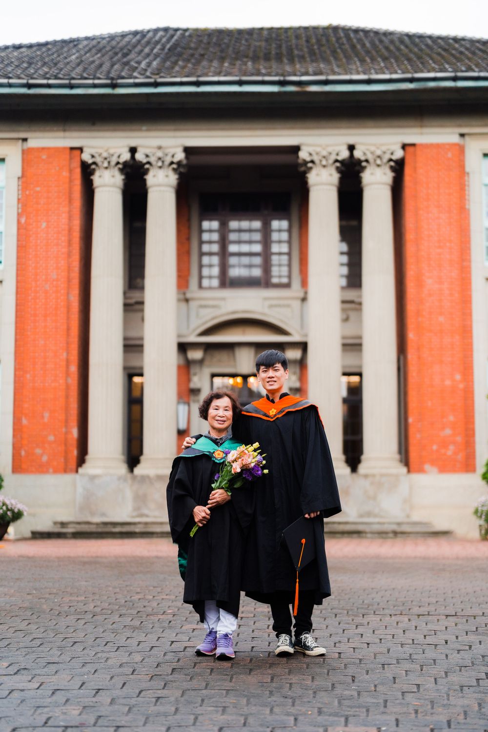 Two graduates pose together in front of orange university building holding graduation flowers.