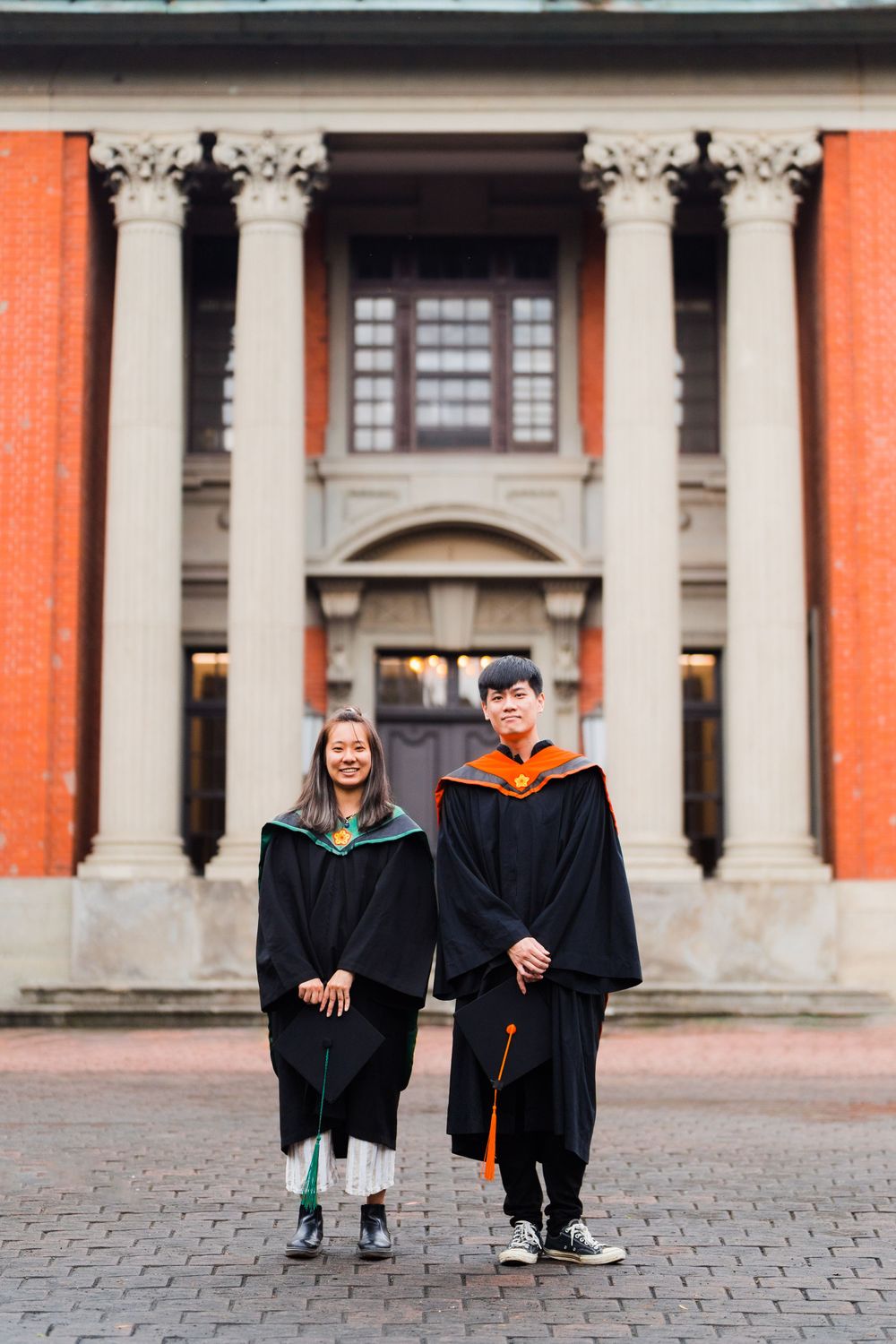 Two graduates in black gowns standing in front of a neoclassical building with orange walls and white columns.
