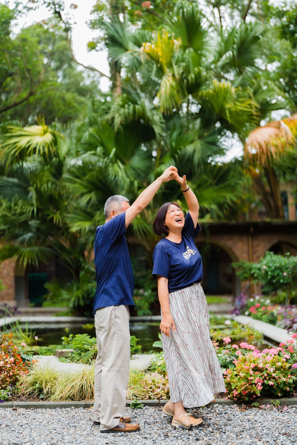 A couple dances playfully in a garden surrounded by palm trees and flowers.