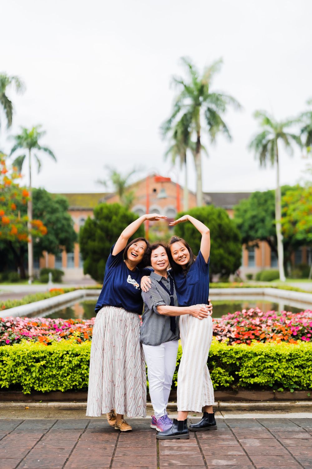 Three friends pose playfully in front of a tropical garden with palm trees and colorful flowers at a university campus.