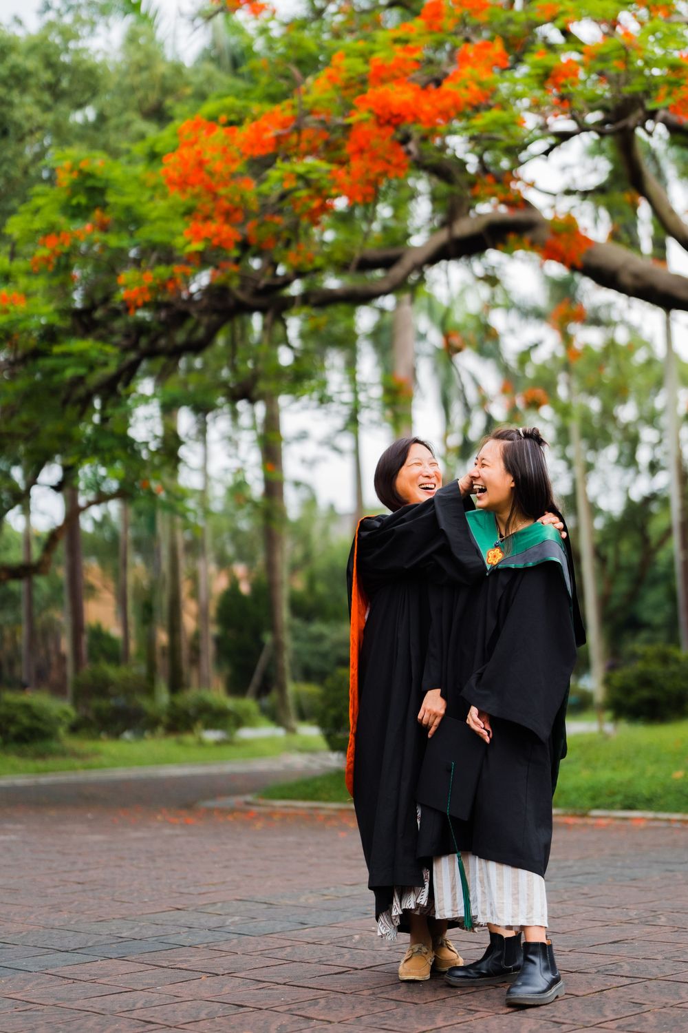 Two graduates embrace and laugh together under orange flowering trees.