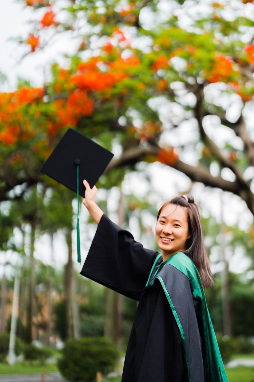 Graduate in black gown and green stole raises cap joyfully under flowering orange trees.