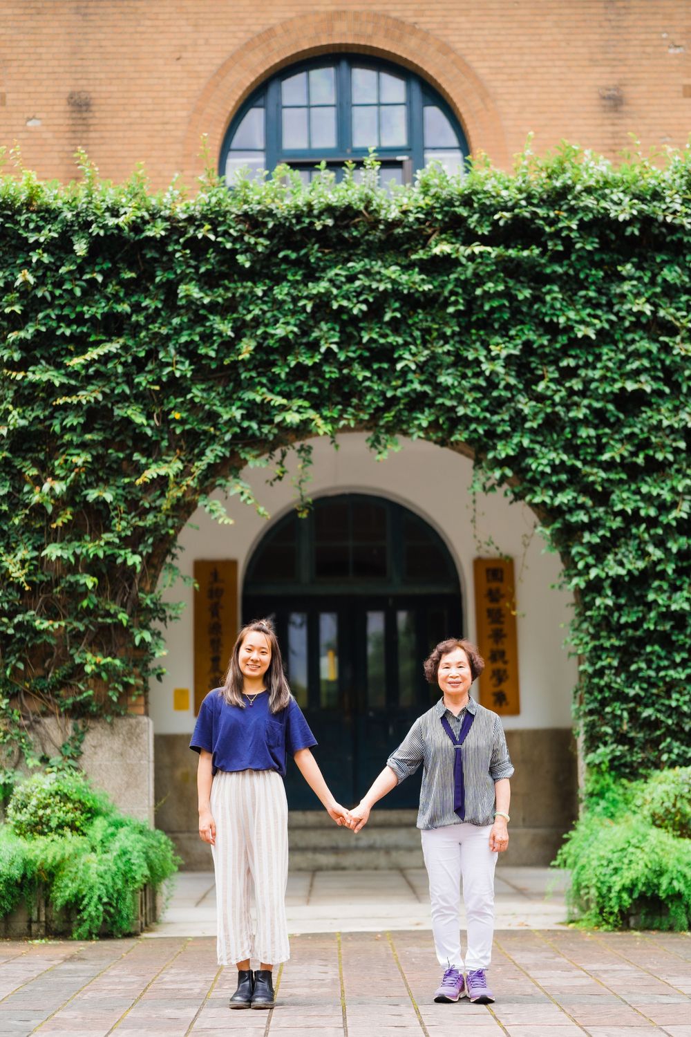 Two people hold hands while standing in front of an ivy-covered building entrance.