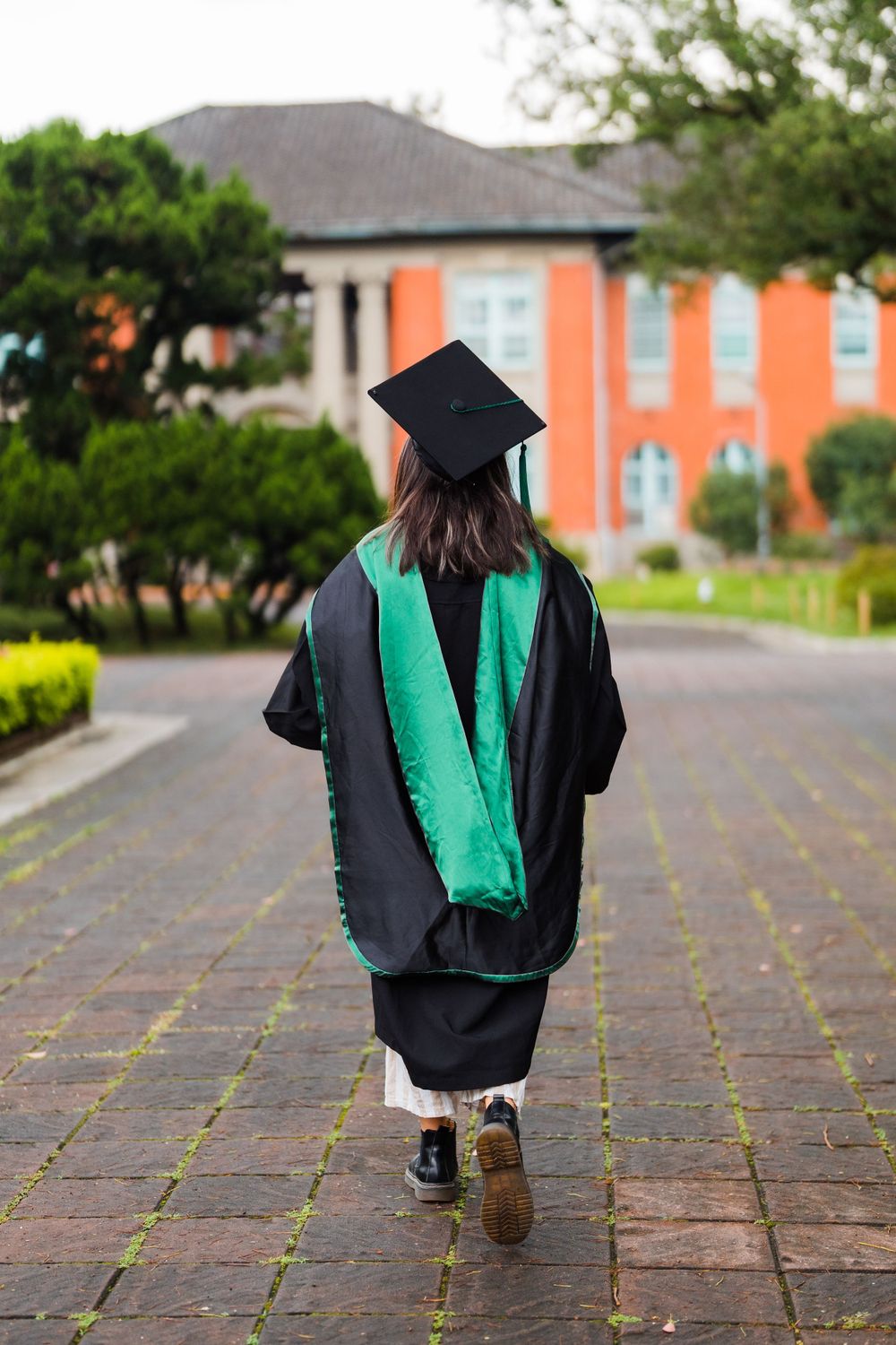 Graduate in black cap and gown with green stole walks away on brick path toward orange building.