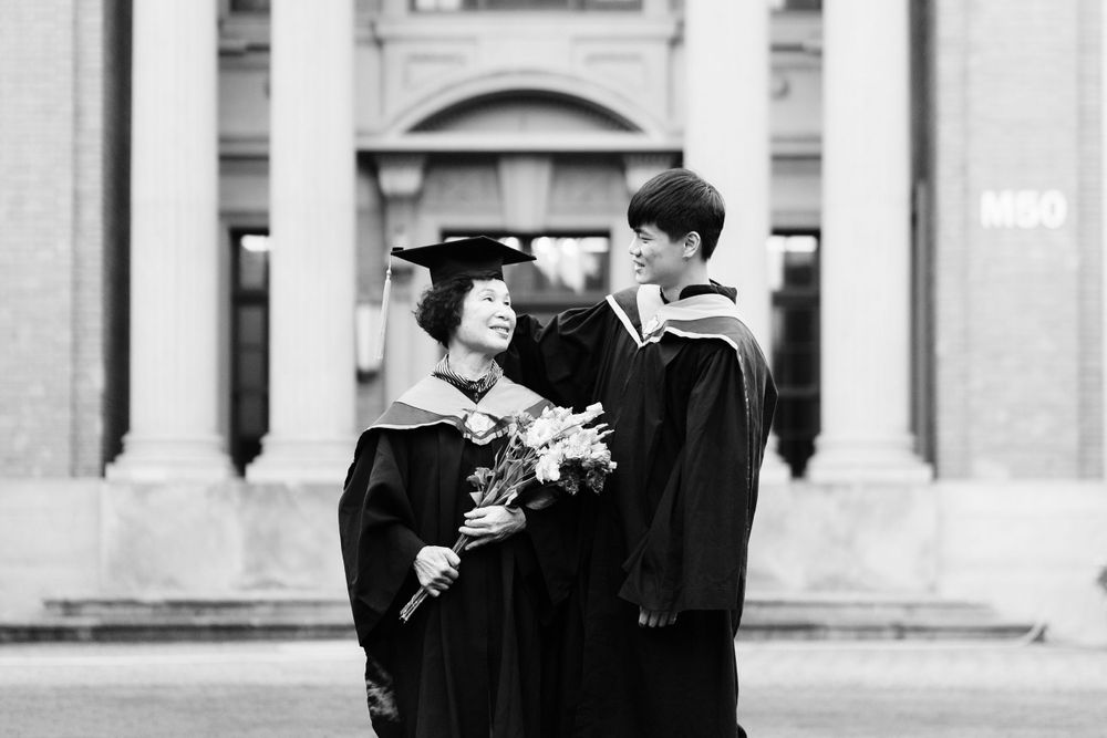 Two graduates in black academic regalia share a joyful moment in front of a classical building.