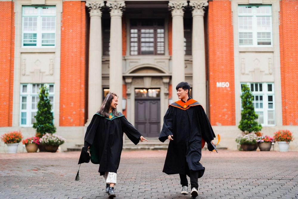 Two graduates in black robes walking joyfully in front of an orange brick building with classical architecture.