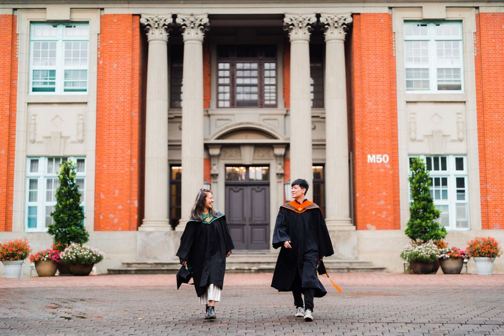 Graduates celebrating their achievement while walking in front of a collegiate building with orange walls.
