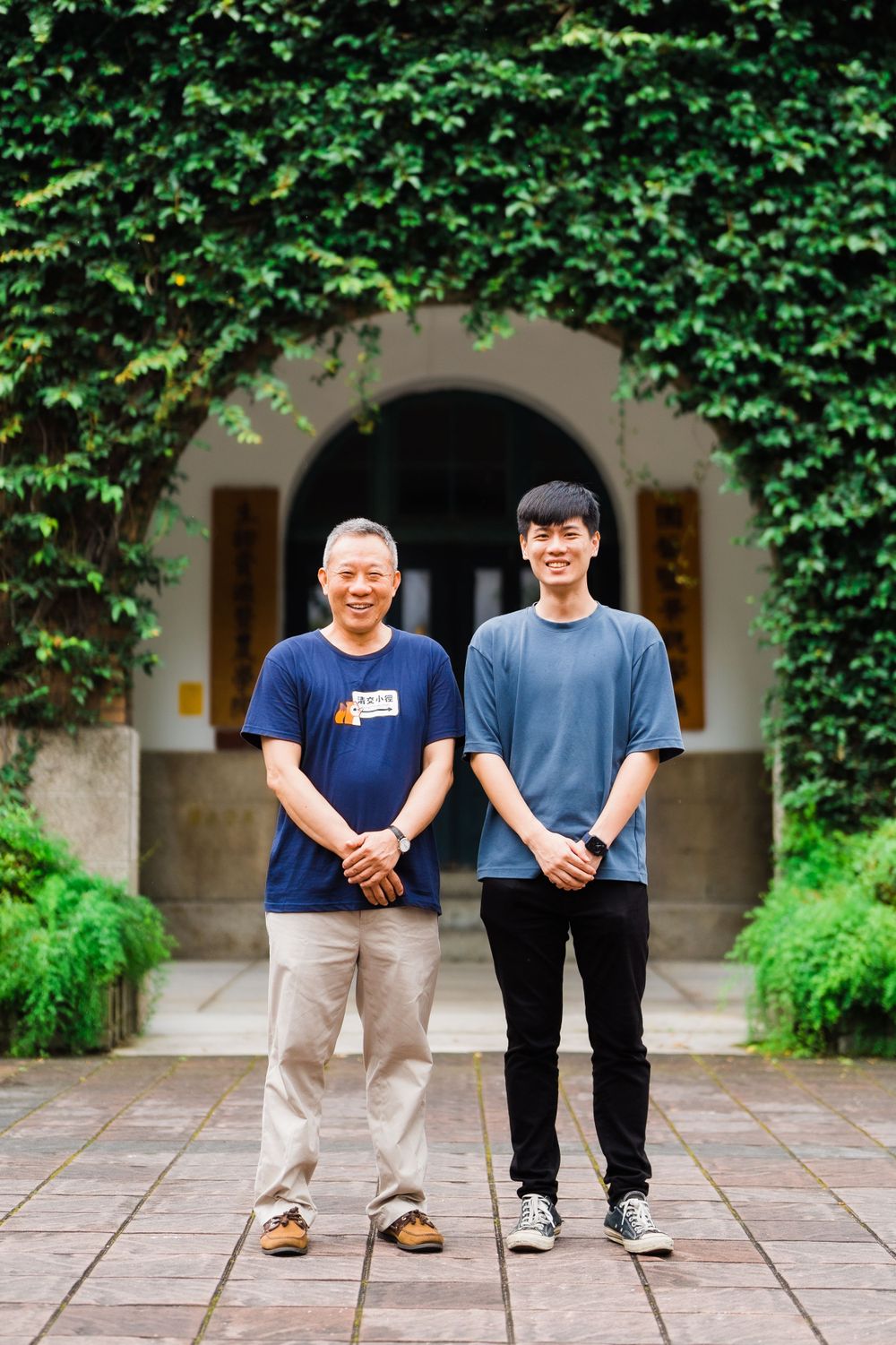 Two people stand together smiling in front of an ivy-covered archway entrance.
