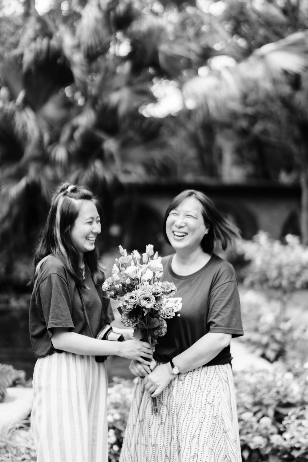 Two friends laugh joyfully while holding a bouquet of flowers in a garden.