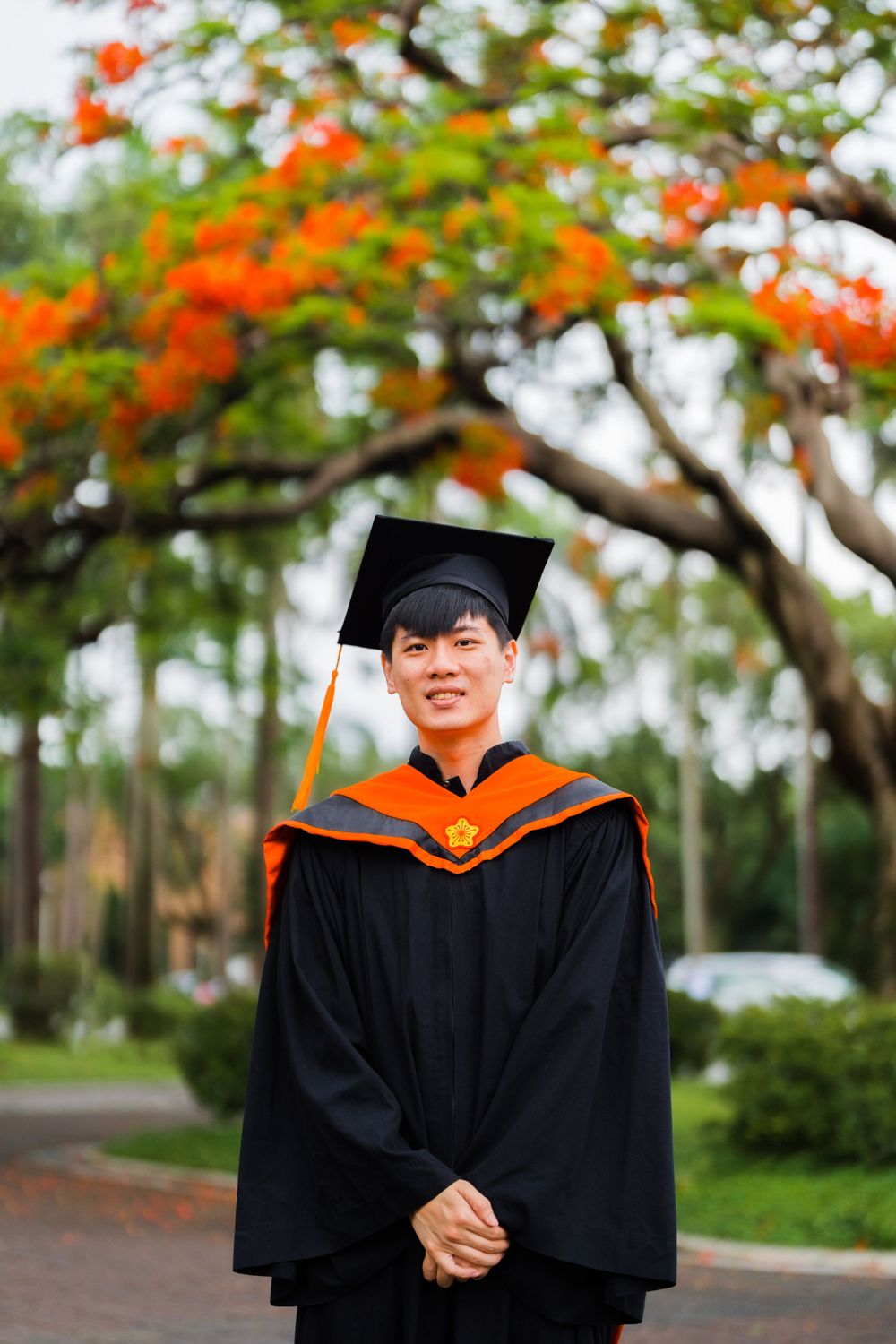 Graduate in orange and black academic regalia poses under flowering tree during graduation photoshoot.