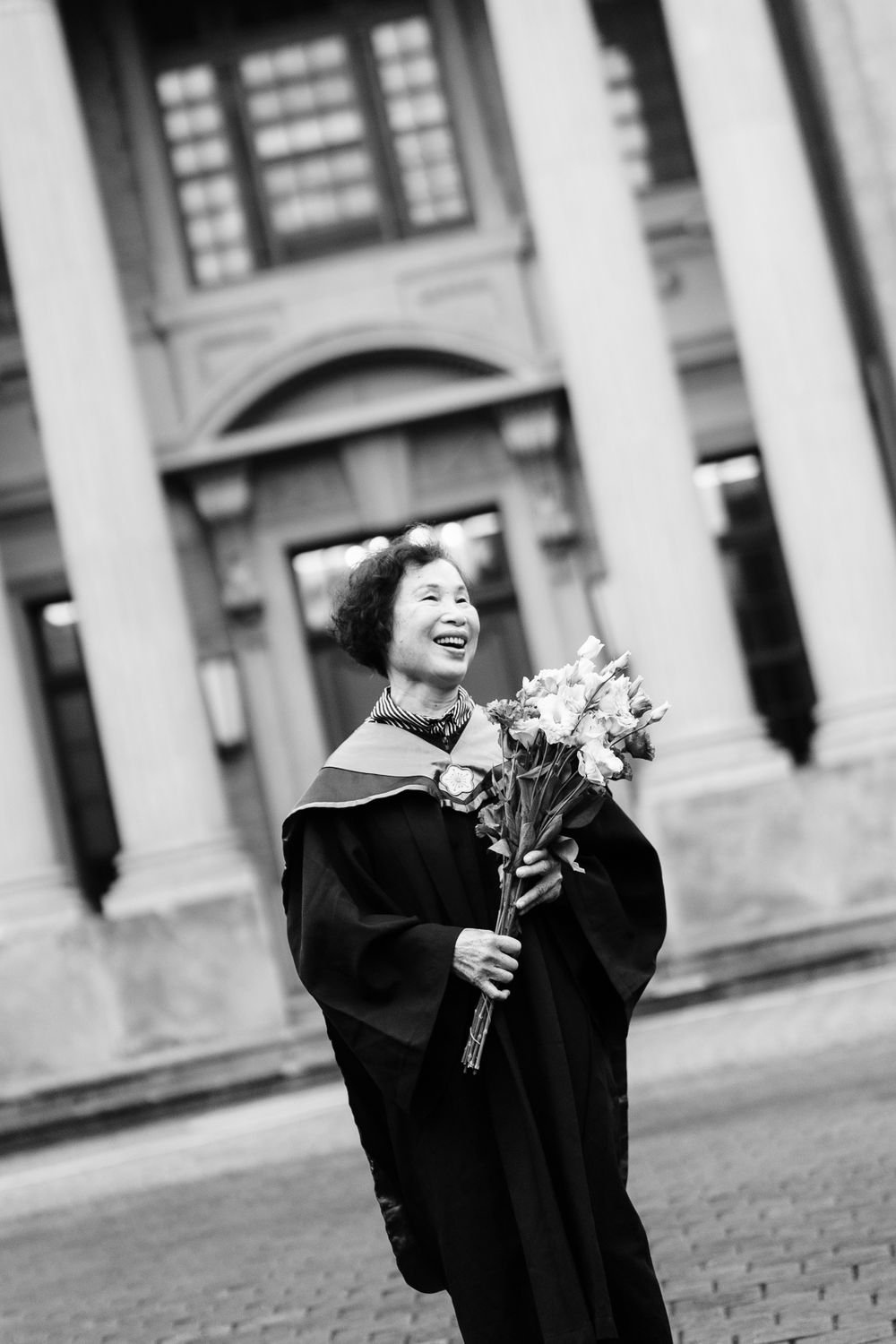 Black and white portrait of graduate laughing while holding bouquet in front of classical building.