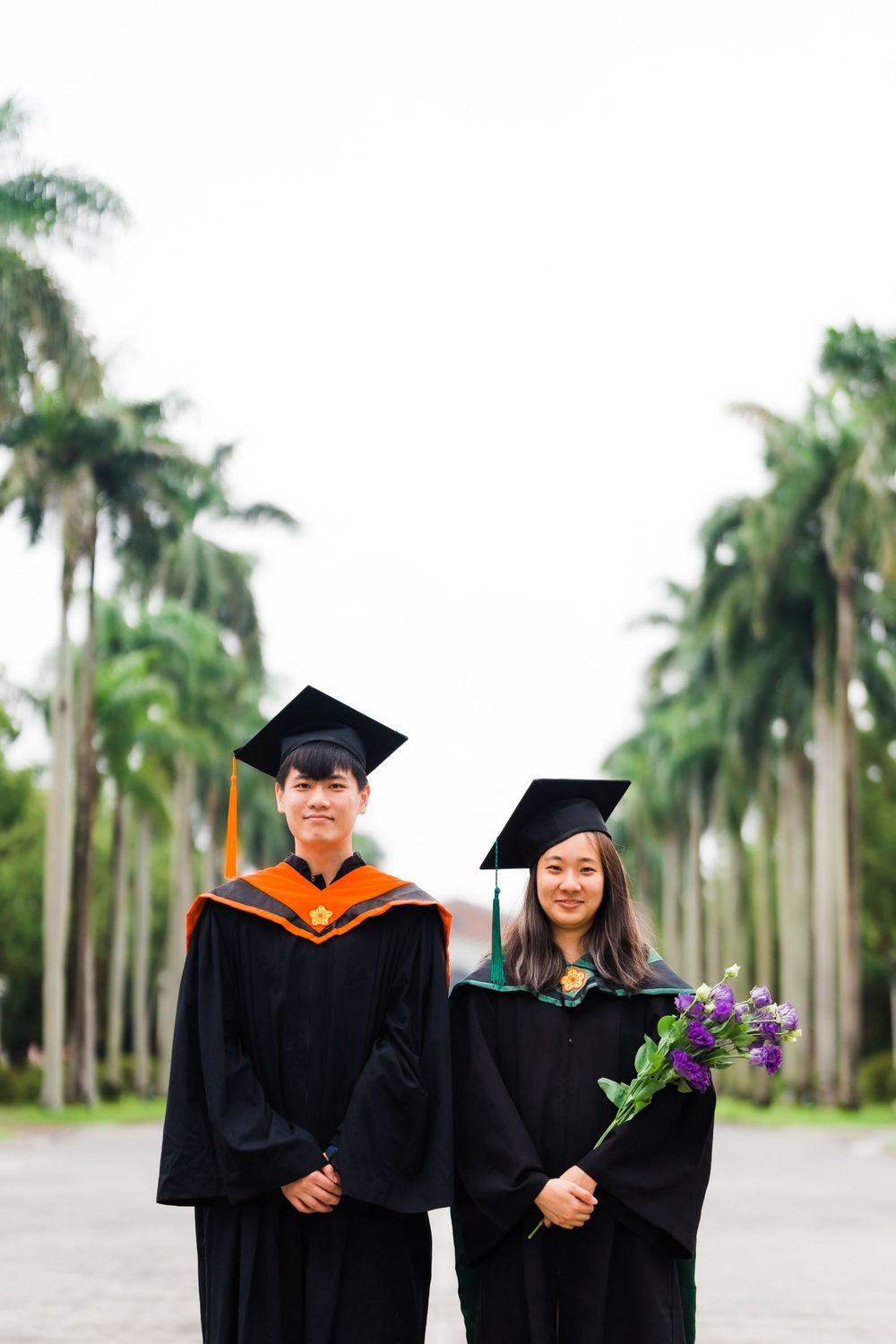 Two graduates in black and orange regalia pose together while holding purple flowers on a palm-lined path.