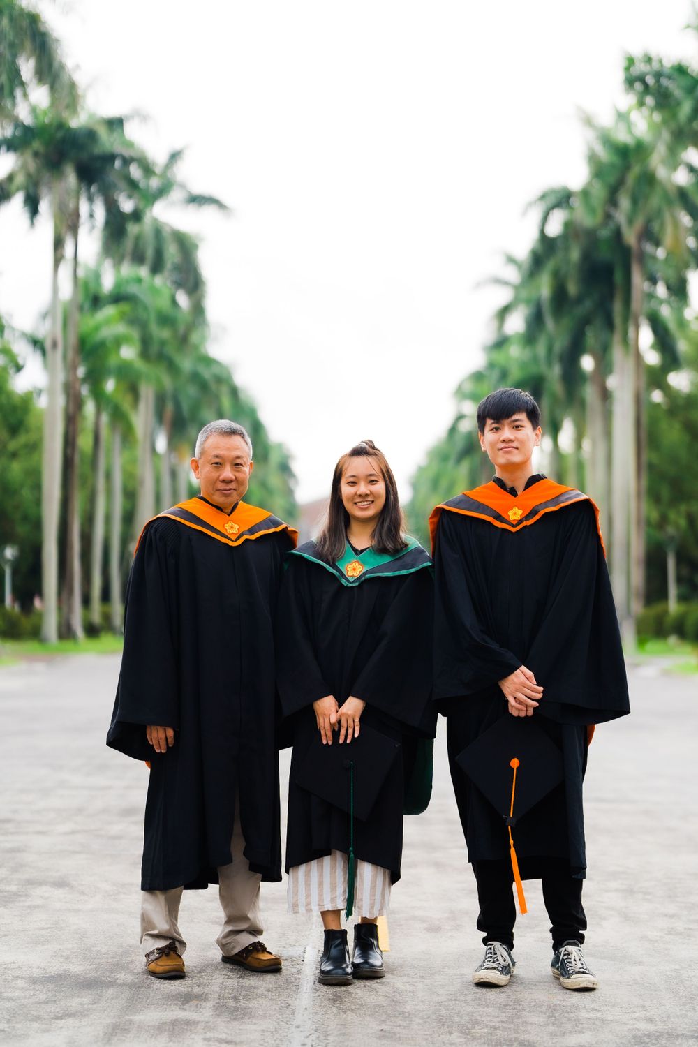 Three people in academic regalia standing on palm-lined path during graduation.