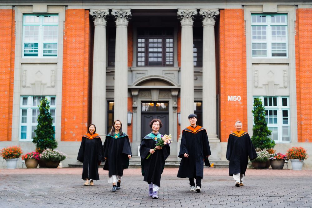 Graduates in black gowns walk together in front of an orange-colored academic building with classic columns.