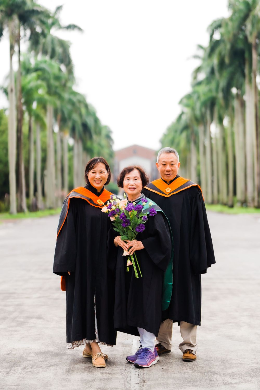 Three graduates wearing academic regalia pose with purple flowers on a palm-lined walkway.