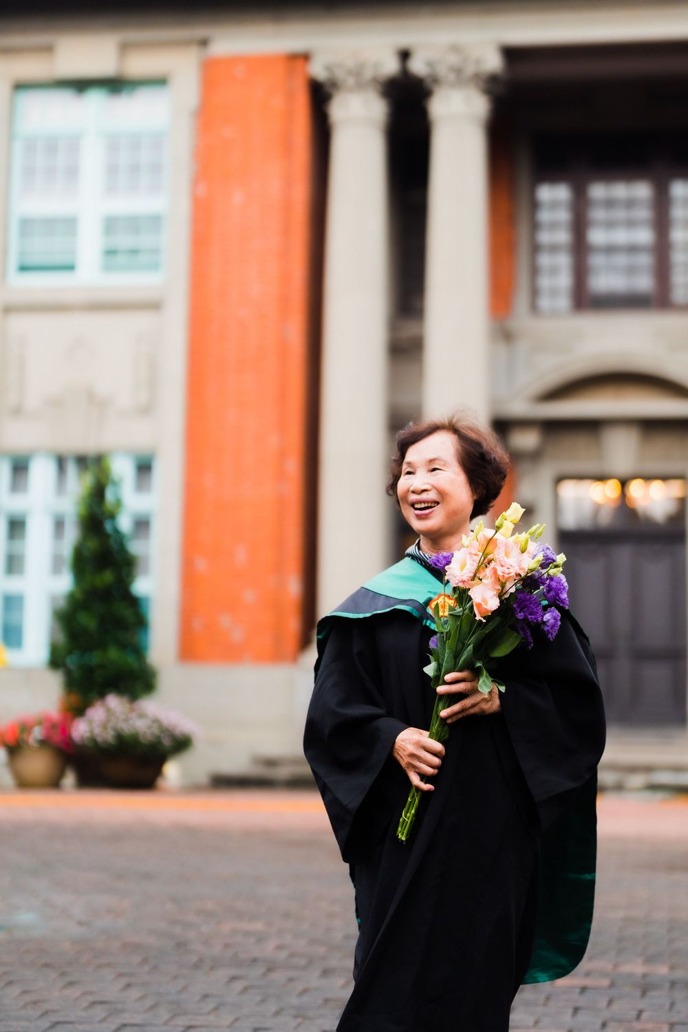 Joyful graduate holding vibrant flowers stands before orange-walled university building.