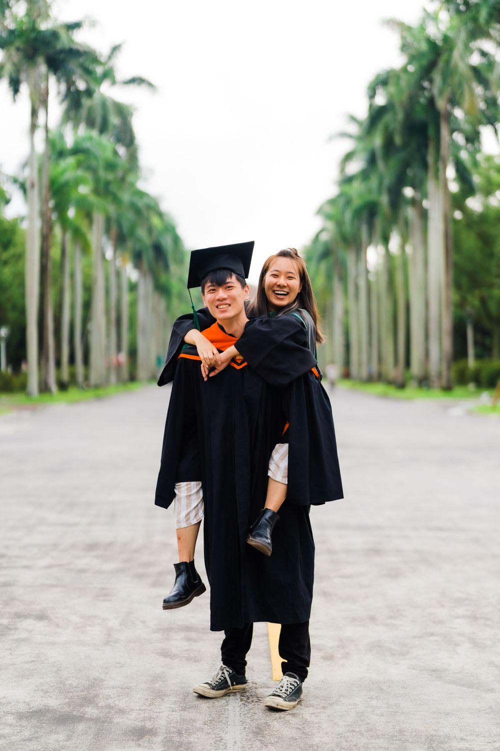 Playful graduation portrait with graduates in black robes against row of palm trees.