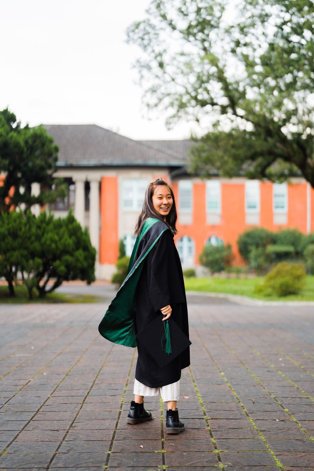 Graduate in black gown and green stole strikes playful pose on brick courtyard.