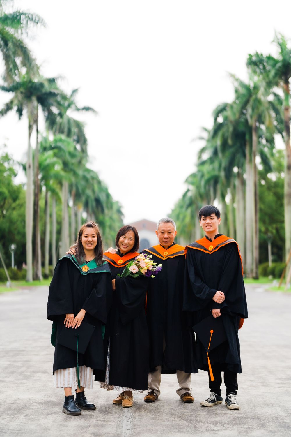Four graduates in academic regalia pose together on palm tree-lined pathway during graduation.