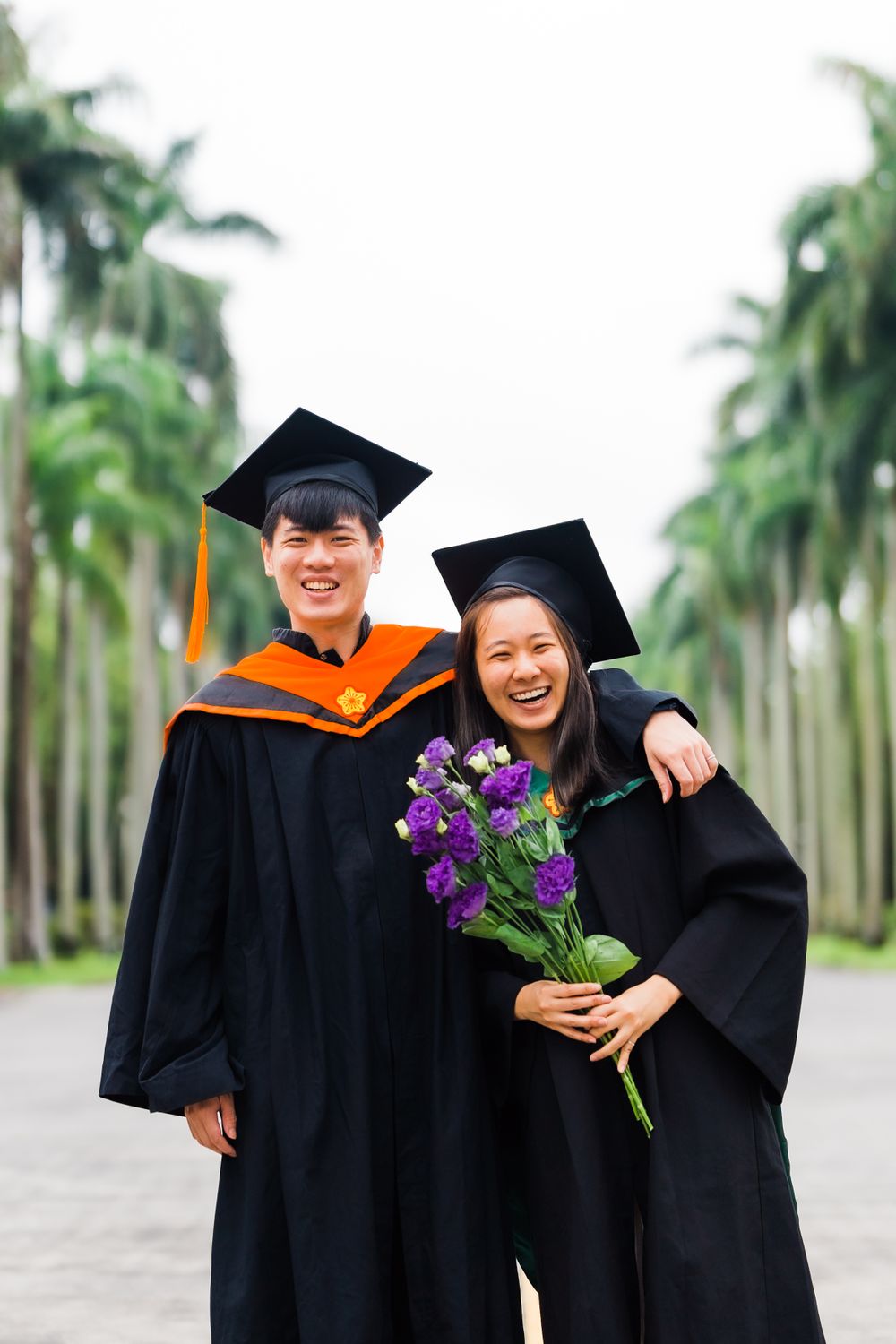 Joyful graduates in academic regalia hold purple flowers while posing between rows of palm trees.