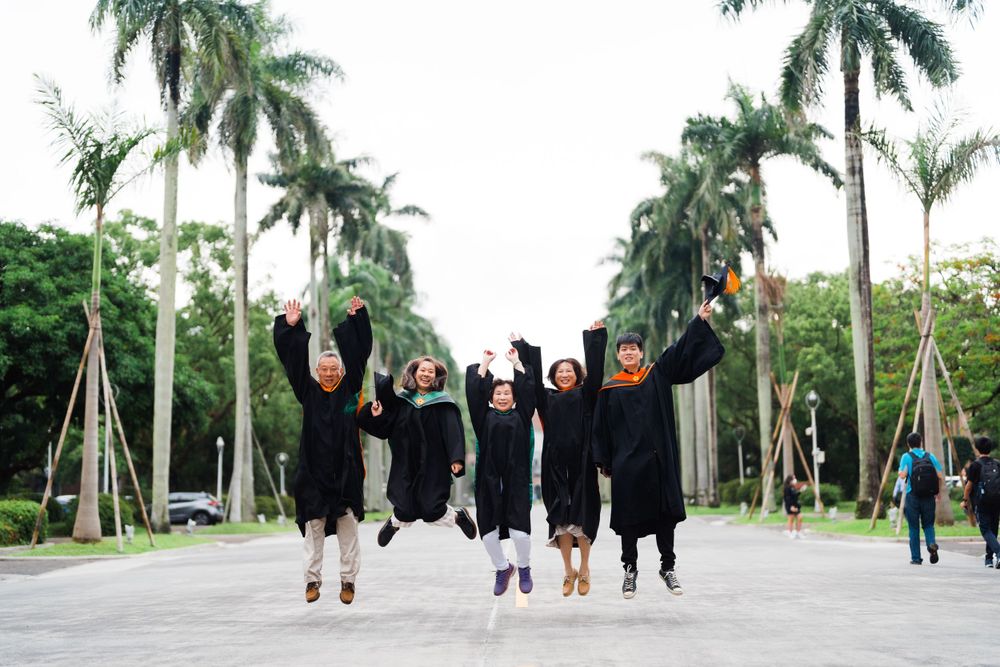 Graduates in black robes jumping for joy on a palm tree-lined path during commencement celebration.