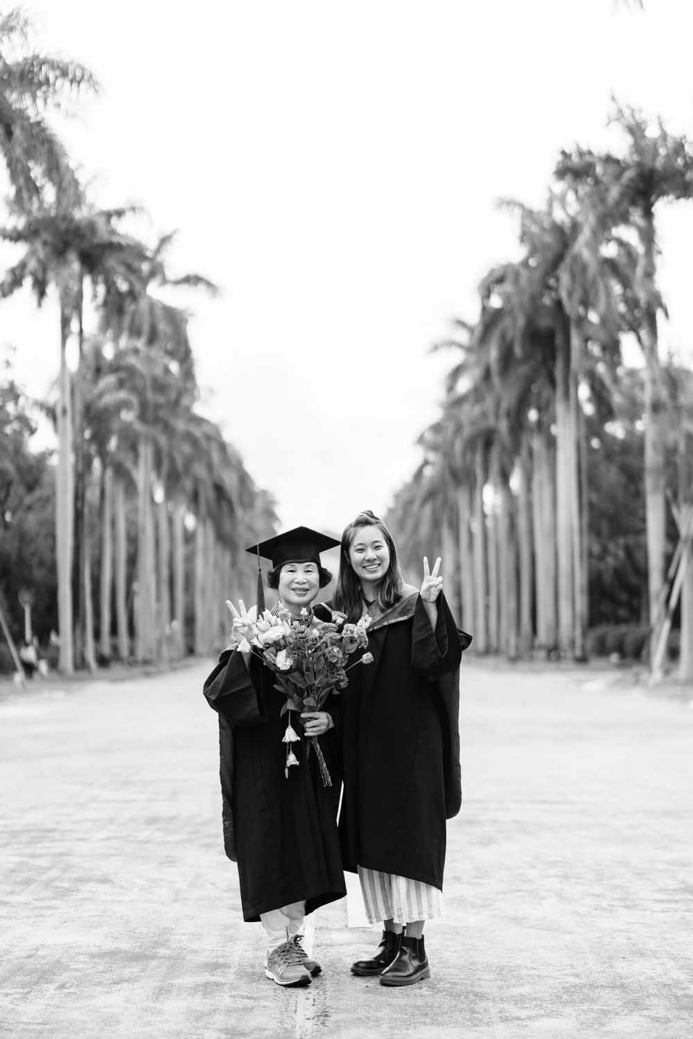Black and white graduation photo of two graduates holding bouquets on a palm tree boulevard.