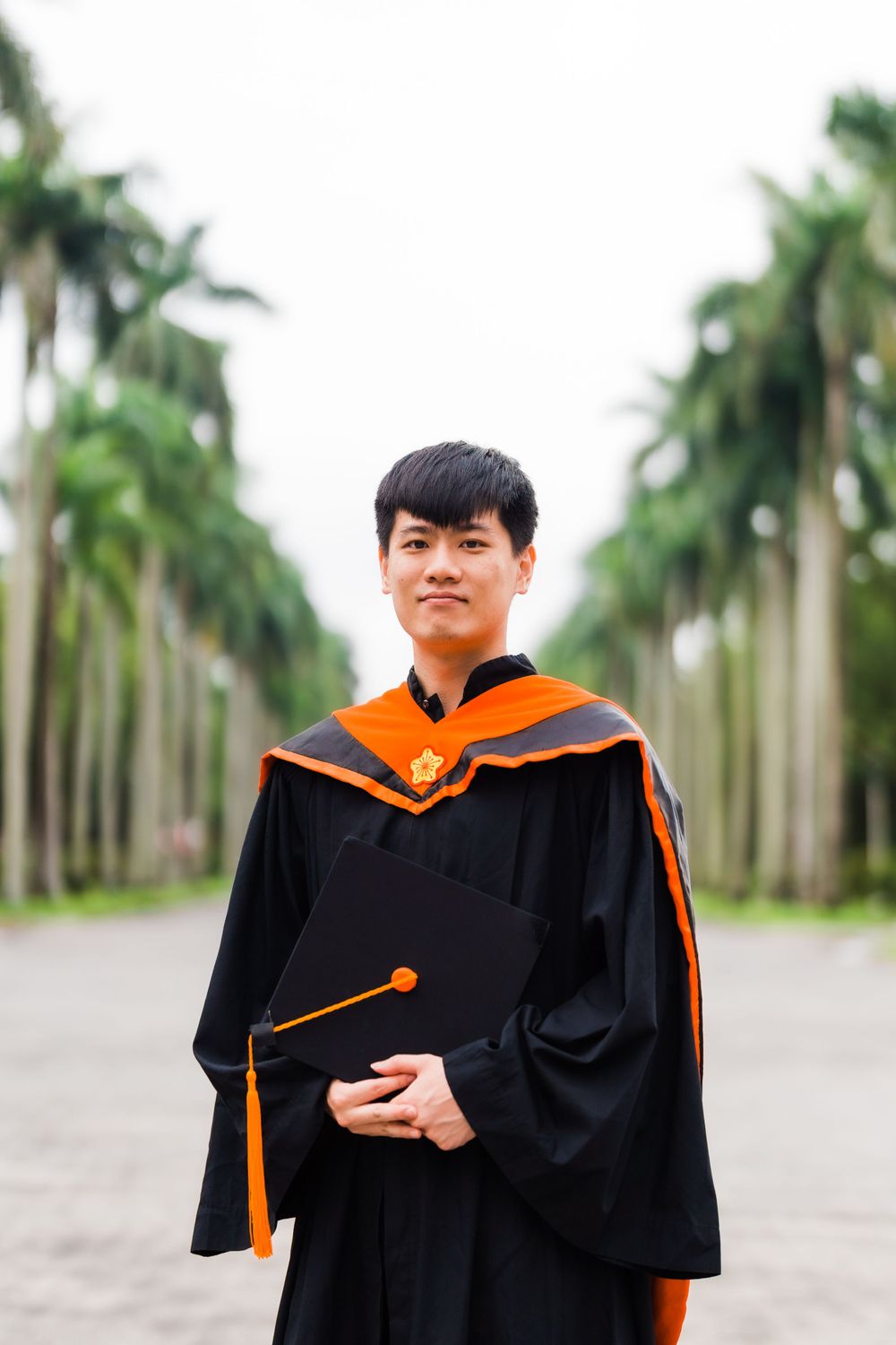 Graduate in black and orange academic regalia stands on palm tree-lined path holding diploma.