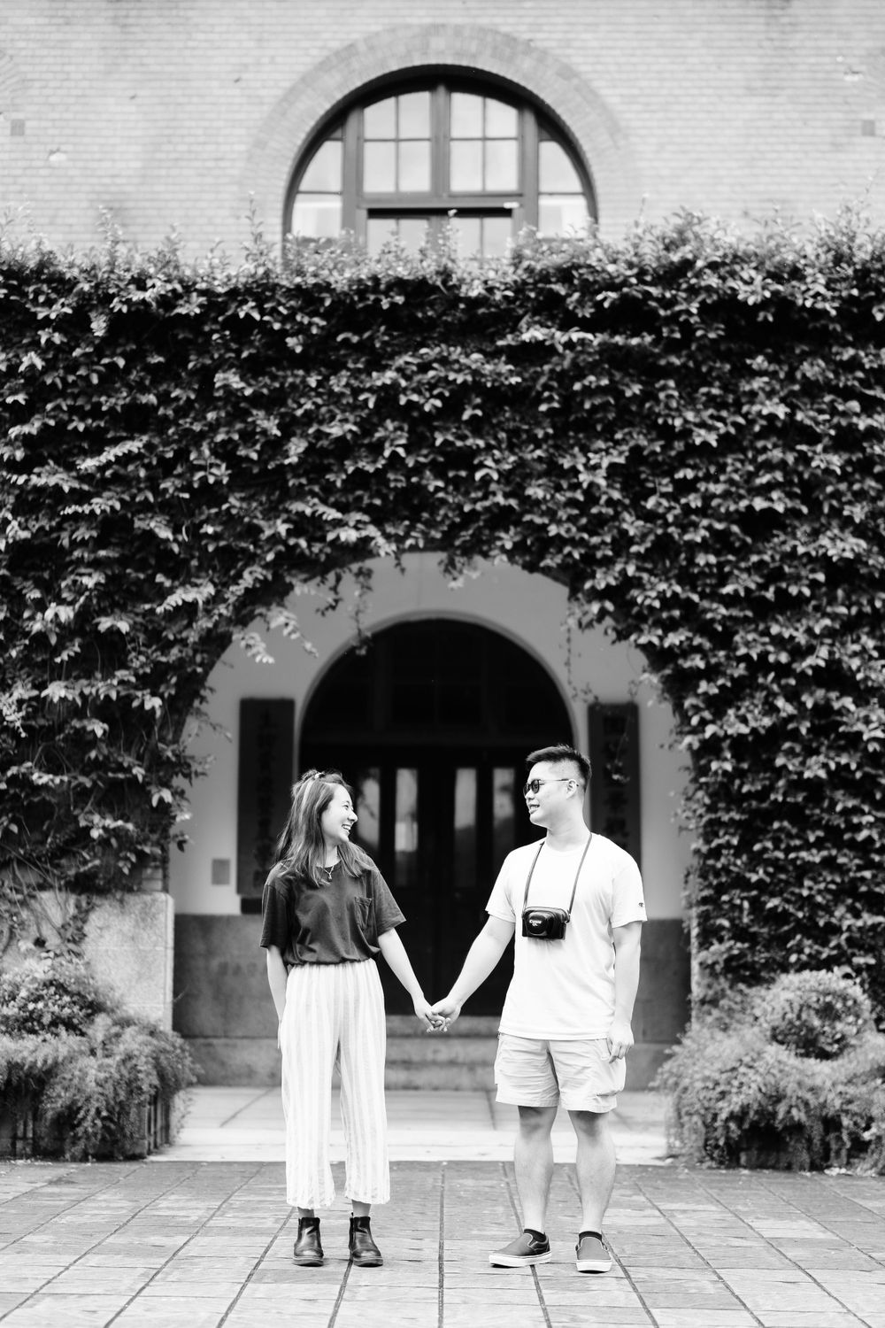 A couple holds hands in front of an ivy-covered building in black and white.