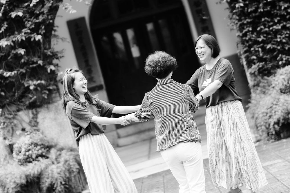 Three people join hands in a circle near a stone archway covered in vines.