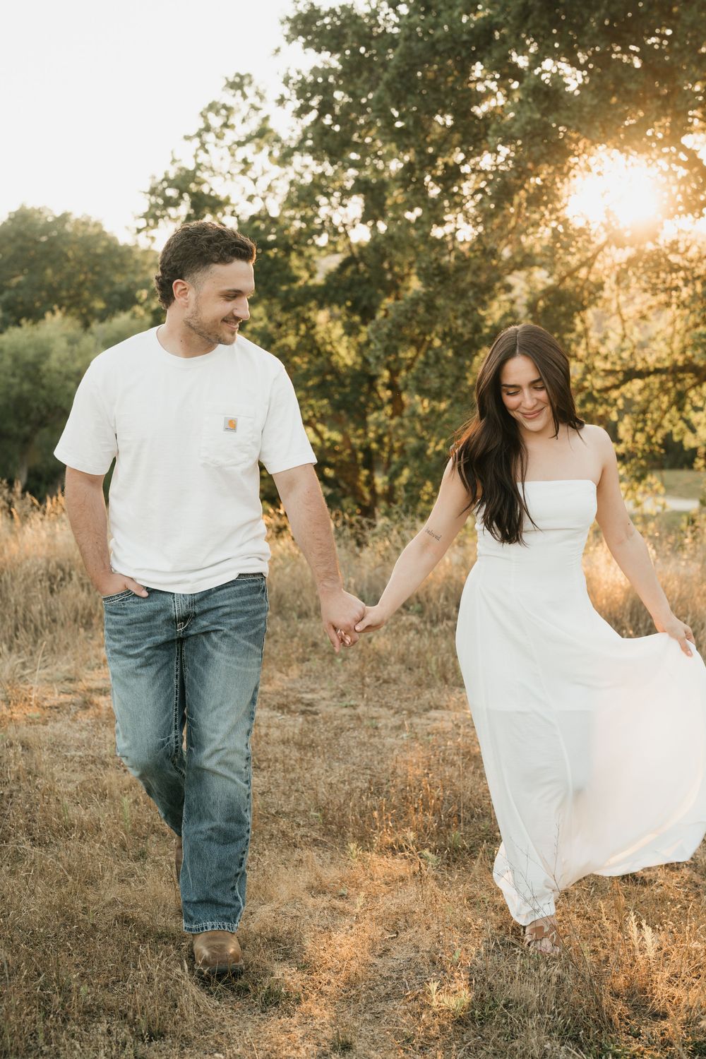 A couple holds hands walking through a golden sunlit field, man in white tee and jeans, woman in flowing white dress.