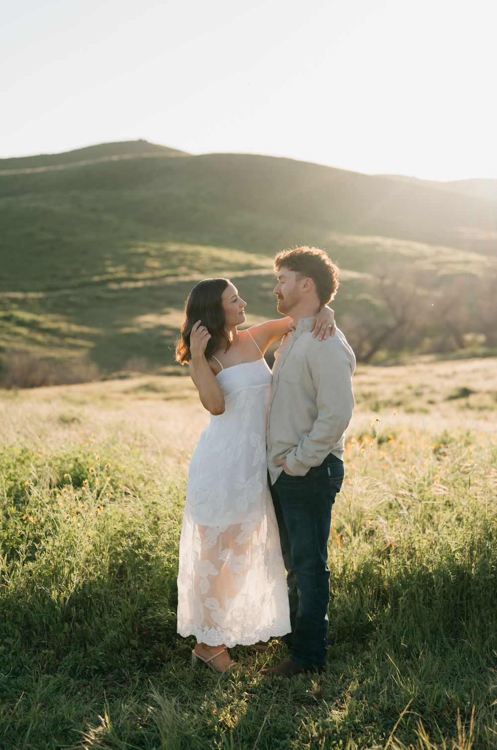 A couple dances together in a sunlit meadow during golden hour, surrounded by rolling green hills and wildflowers.