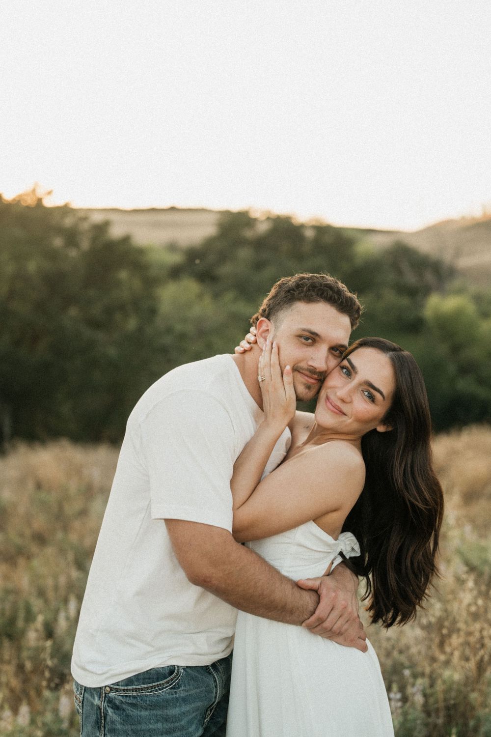 Couple embracing in a golden field at sunset, woman in white dress touching man's face, trees in background.