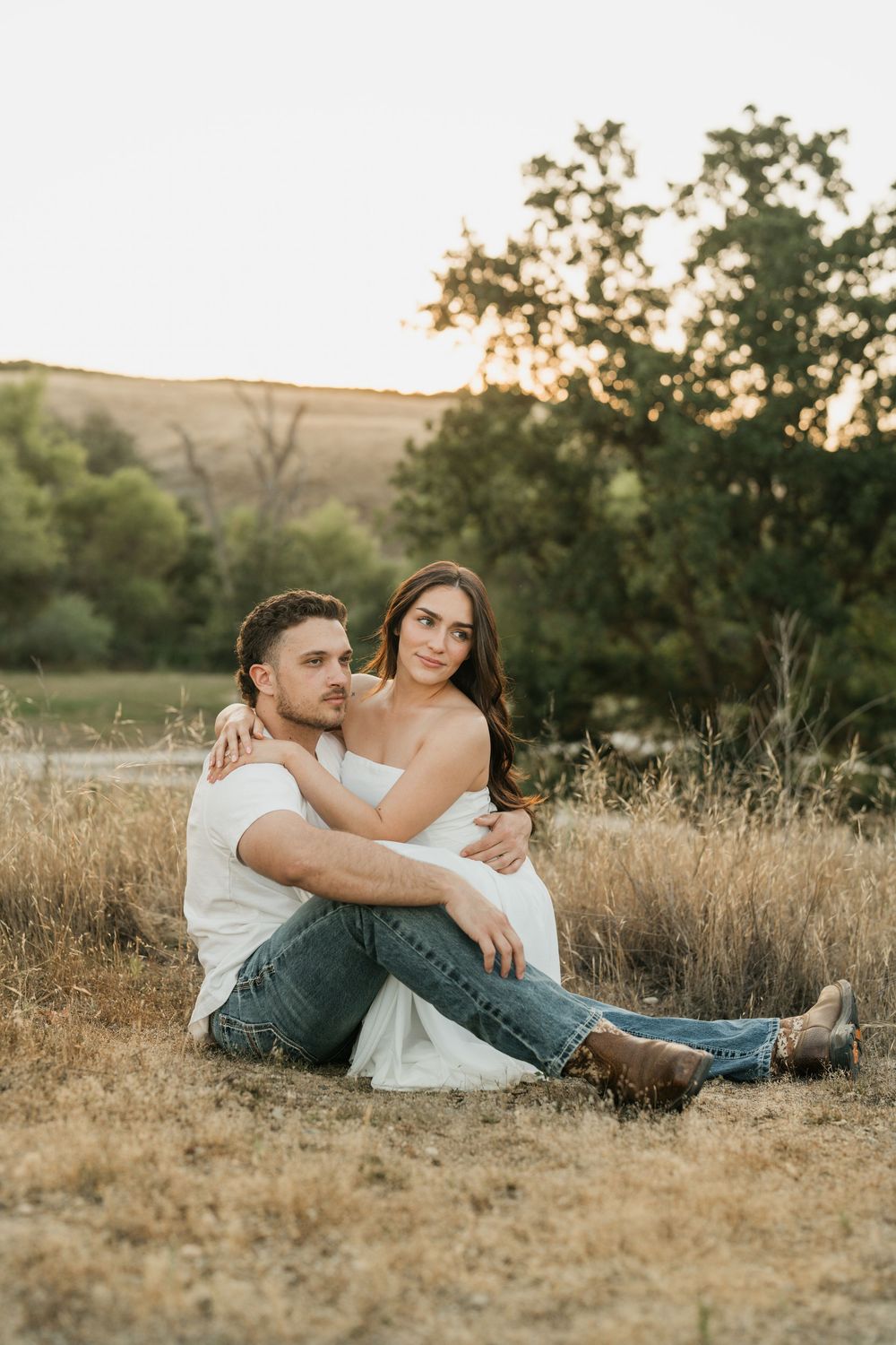 A couple sits together in a golden field at sunset, the woman in white embracing the man in jeans and a white tee.