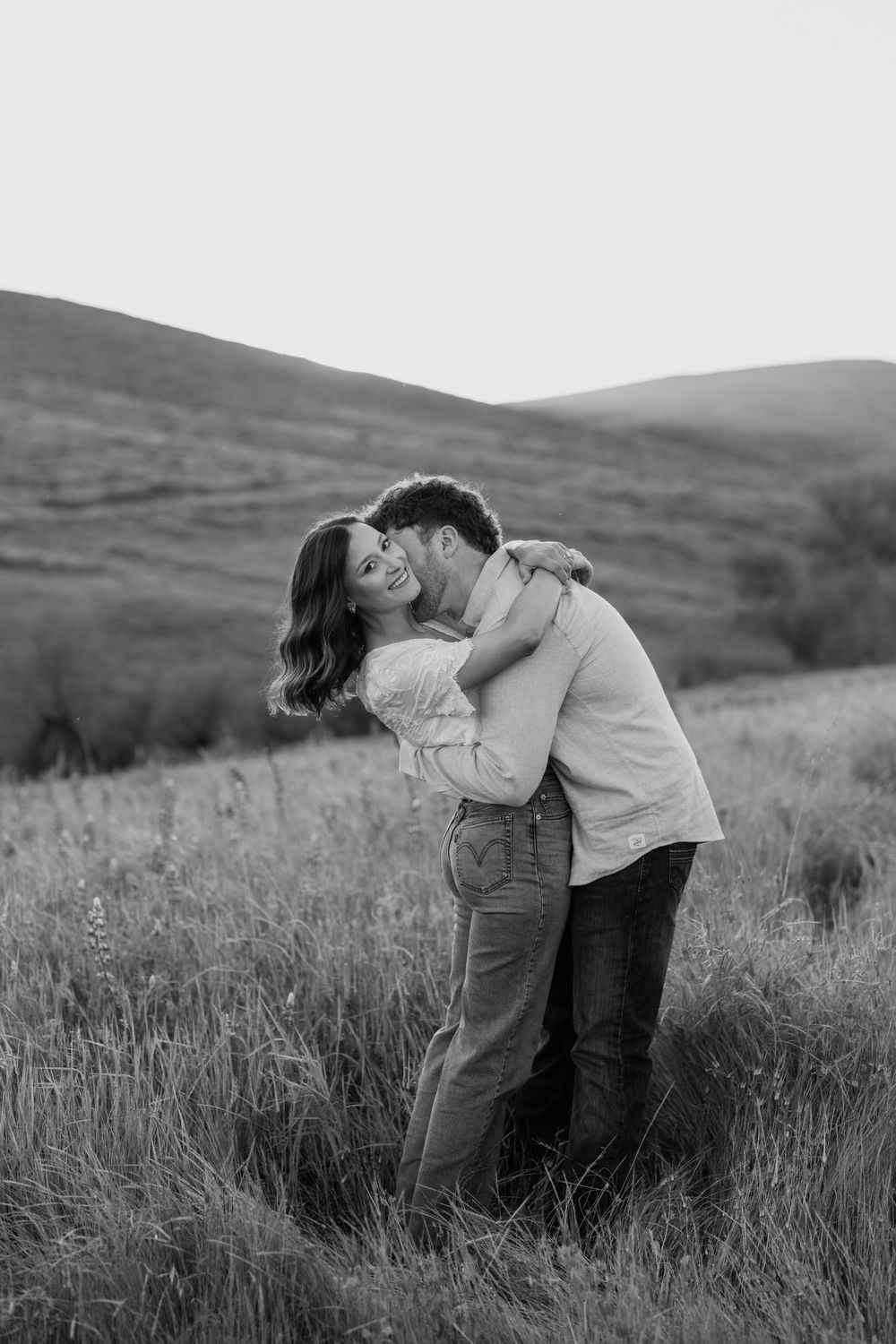 A couple shares a romantic kiss in a grassy field with rolling hills in the background, captured in black and white.