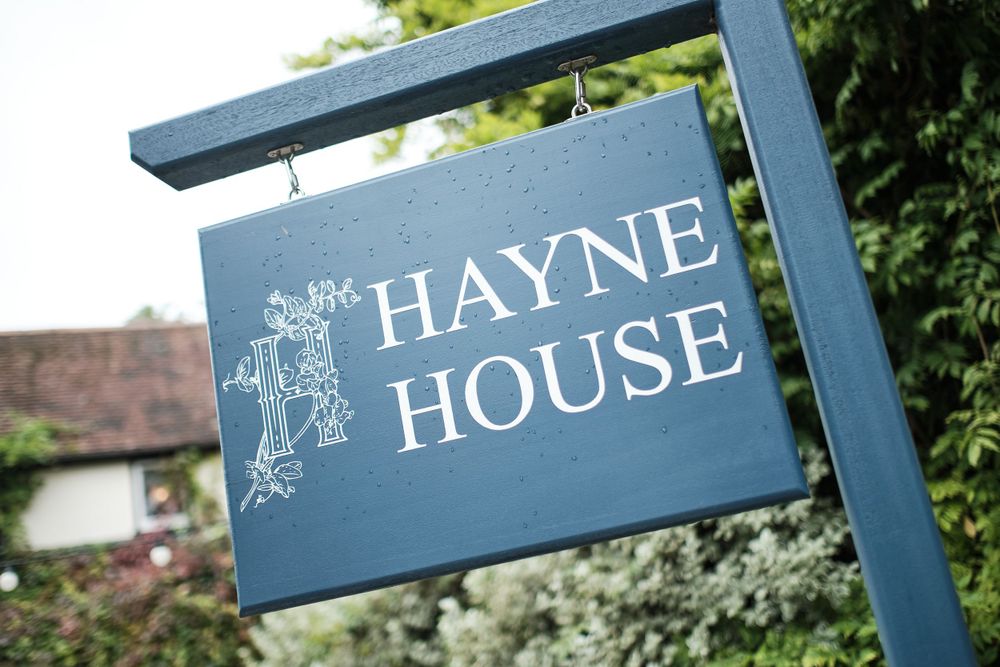 Blue wooden sign for Hayne House mounted on post against garden background.