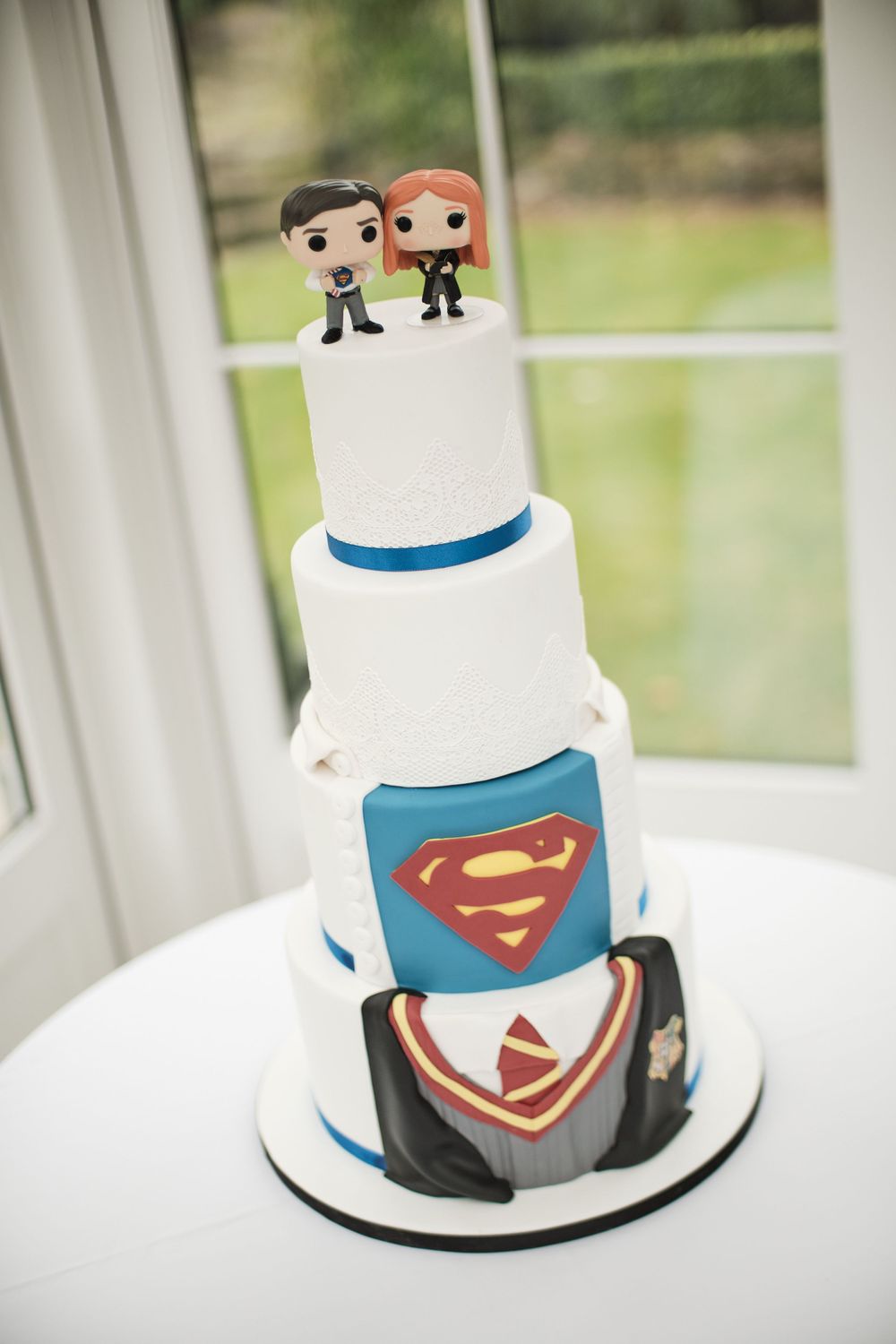 Three-tiered white wedding cake decorated with Superman logo and Funko Pop figurine toppers.