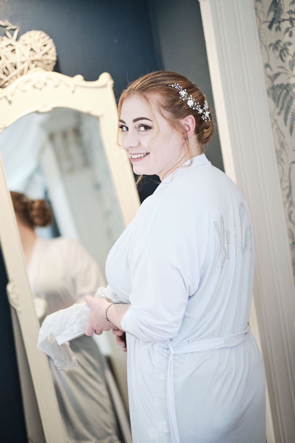 Bride in white robe smiles while getting ready in elegant dressing room with ornate mirror.