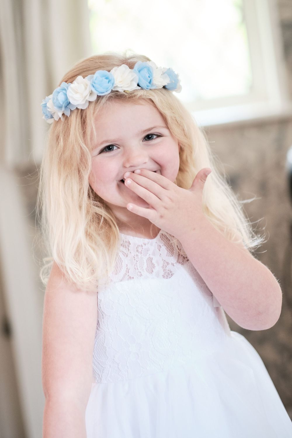 Young flower girl in white dress wearing blue and white floral crown giggles with joy.