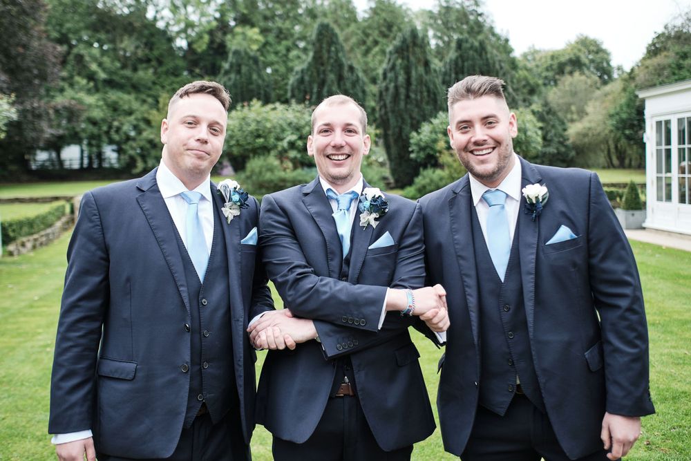 Three groomsmen in navy suits share a laugh on manicured garden grounds.