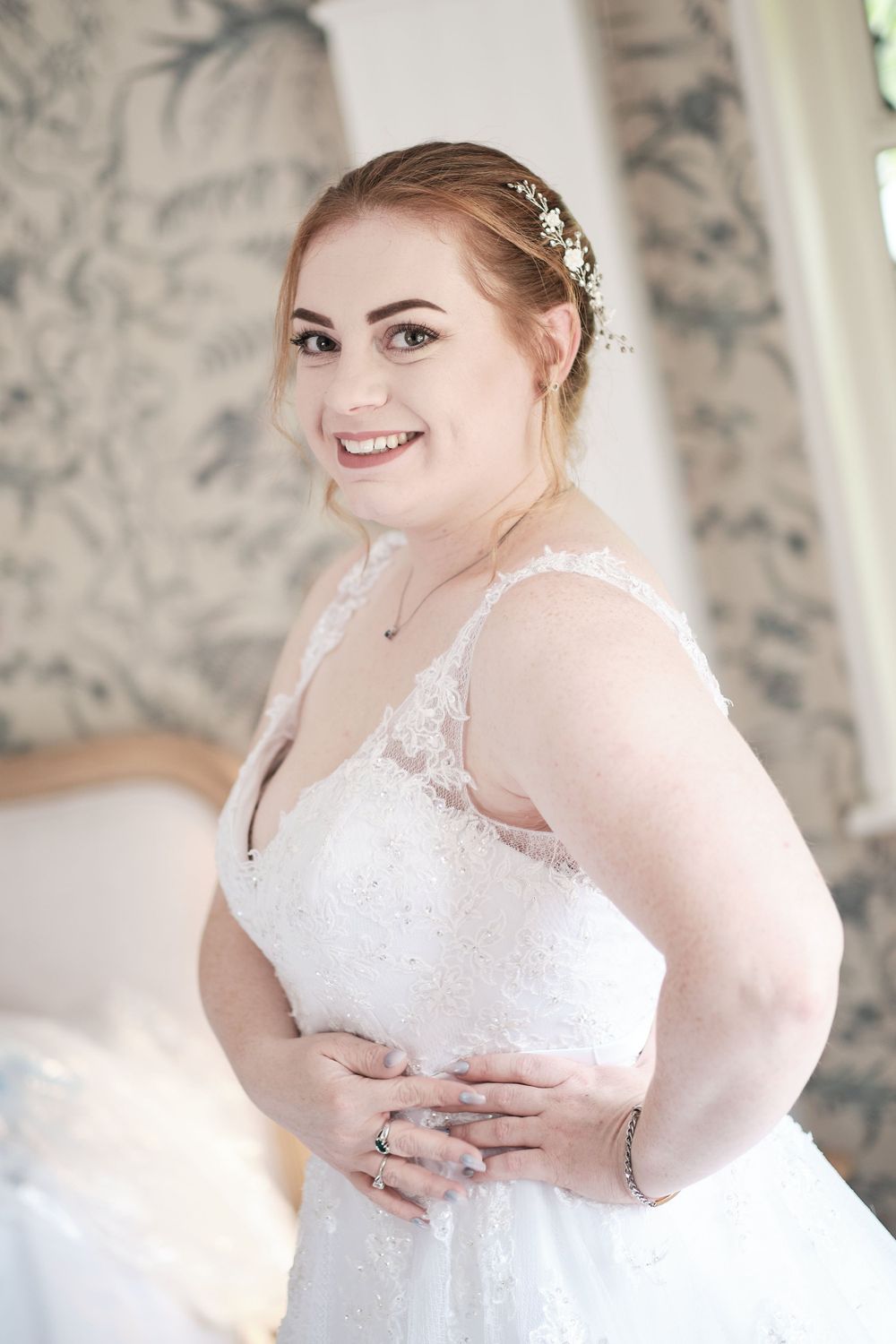 Bride in lace wedding gown shows off her ring while posing in elegant room.