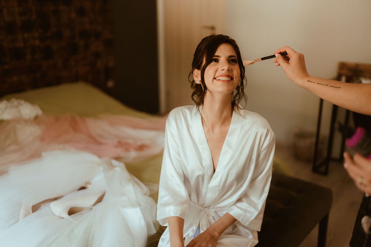 Bride in white silk robe getting makeup done during wedding preparation.