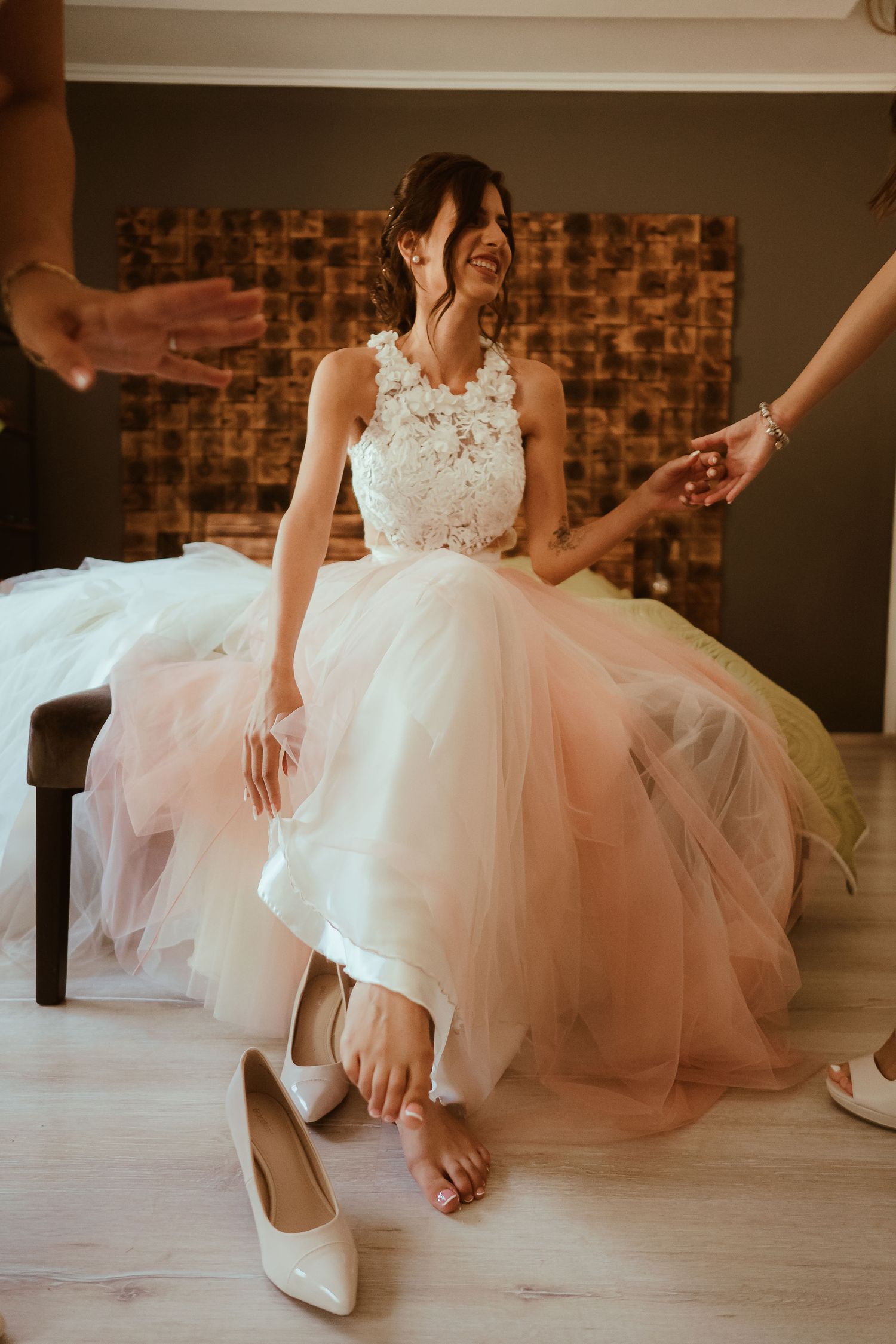 A bride in a white and pink tulle wedding dress joyfully reaches towards white heels placed on a light floor.
