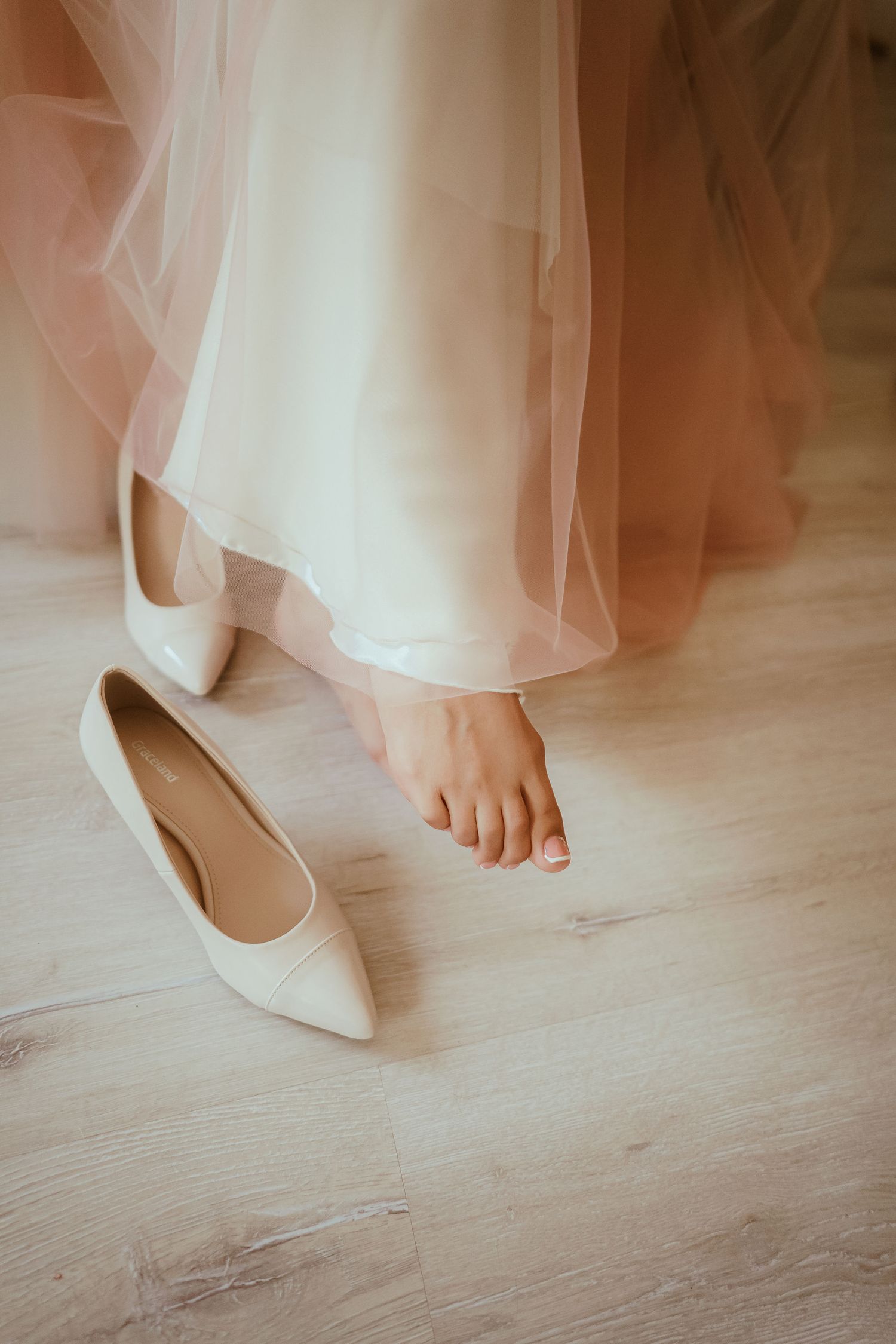 Close-up of white bridal shoes next to bare feet peeking beneath a tulle wedding dress.