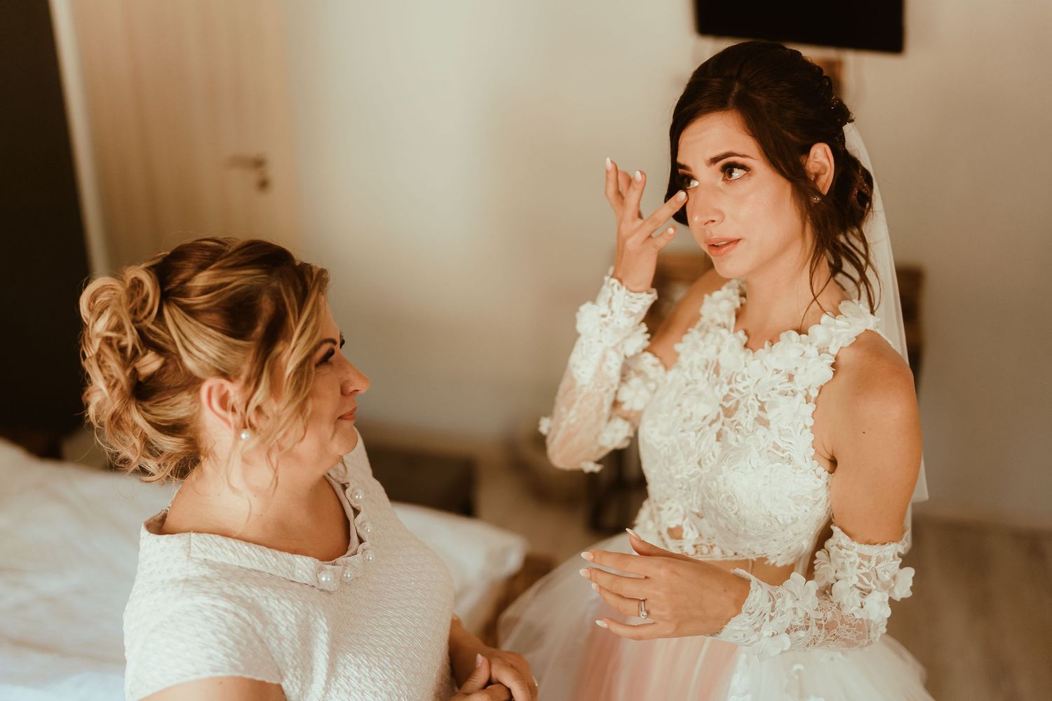 Two women in white lace dresses share a playful moment during wedding preparations.