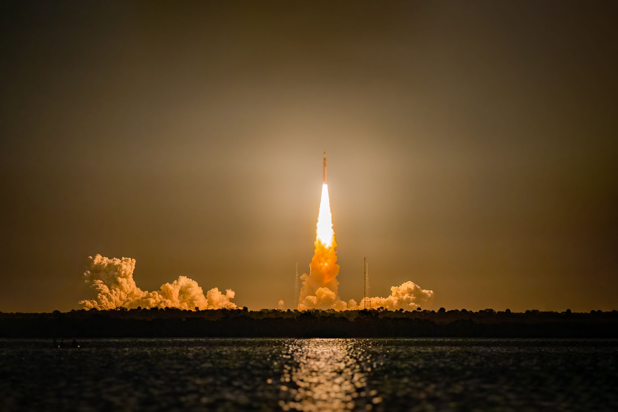 A rocket launch at night creating a bright orange glow reflected on water below.