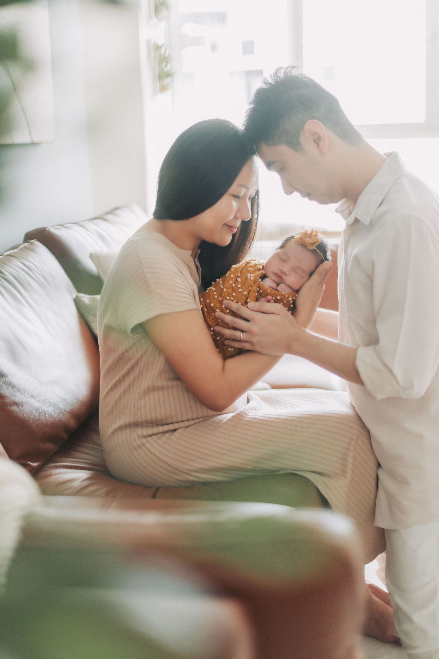 Young Asian parents sitting on sofa gazing lovingly at their newborn baby in bright natural light.