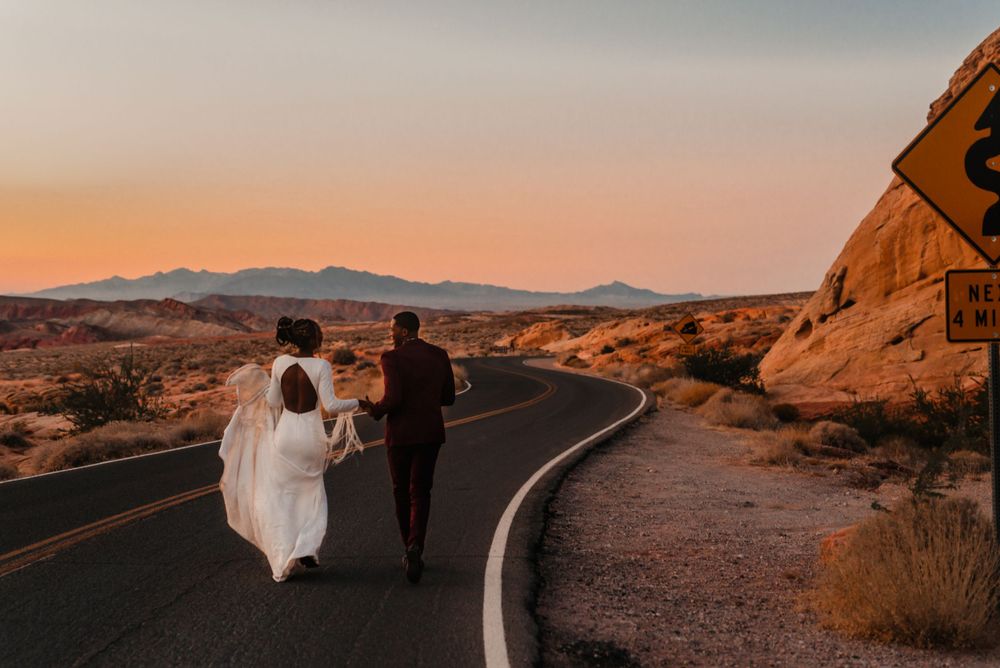A wedding couple walks along a winding desert road during sunset with red rock formations in the background.