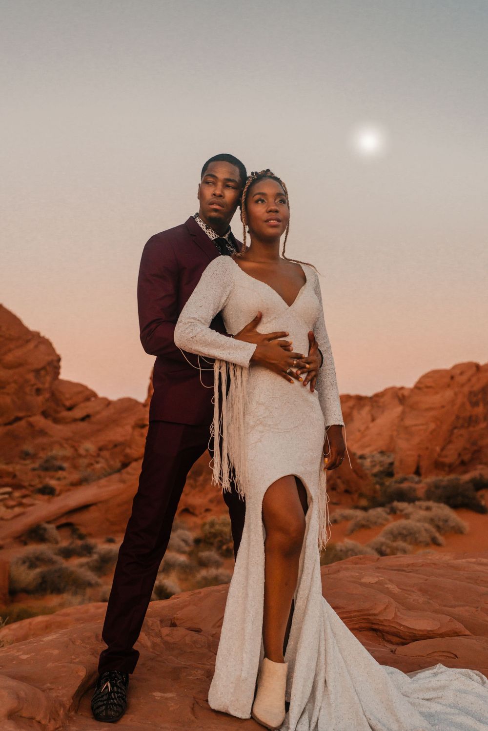 A couple in formal attire poses against red desert rocks during golden hour.