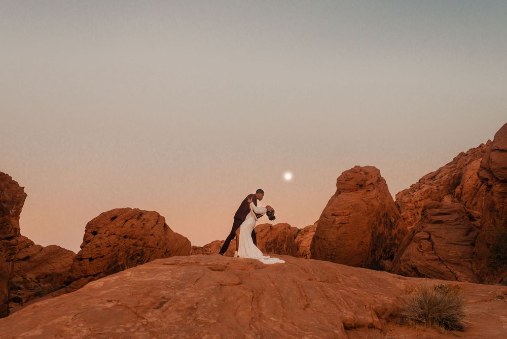 Silhouetted wedding couple stands atop red rock formation at dusk.
